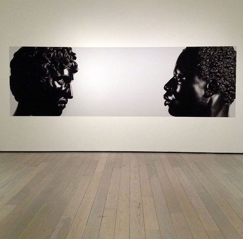 Photograph. From opposite ends of a long composition, two black stone busts face each other. To our left, Antico’s sculpture shows a person with full, curly hair, a straight nose, a moustache, and defined cheekbones. His chin is slightly tucked in. To our right, the face of Harwood’s sculpture seems to look levelly back. It depicts a Black man with short, curly hair, a softly curved nose, and full lips which are closed. Both are shown to the tops of their shoulders.
