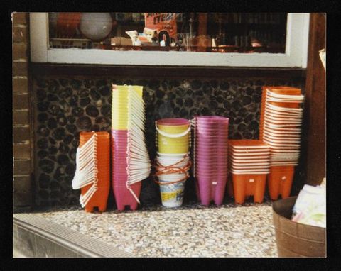Colour photograph. Buckets and moulds for making sandcastles are stacked in six columns. Most of the moulds and buckets are orange, pink, or yellow, all with white handles. Some of the buckets are printed with what could be a landscape scene, and those have orange handles. They seem to be collected on a countertop.