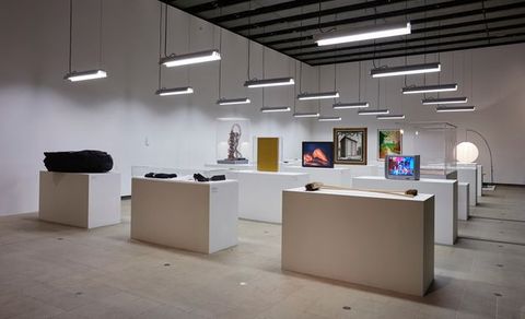 Installation. About a dozen long tables create rows across a long, white, nave-like room. Fluorescent lights hang down over the tables. Objects displayed on the tables include brooms, a pair of shoes, a television playing a video, and at least one photograph.