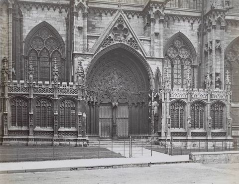 Black and white photograph. A double door on one porch of a cathedral is engulfed within nested arches, which are then crowned by a pointed pediment. Windows, buttresses, and rows of spiky pinnacles are heavily ornamented.