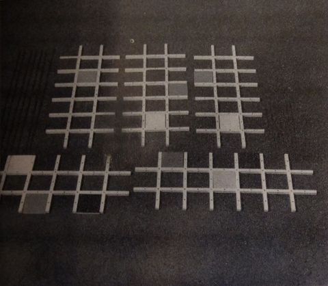 Sculpture. Six lattices, like pieces of a trellis, lie against a dark ground. Each is made of two long rows overlapping six shorter rows, like an elongated tic-tac-toe board. Each piece has one, two, or three cells filled in with solid squares in varying shades.