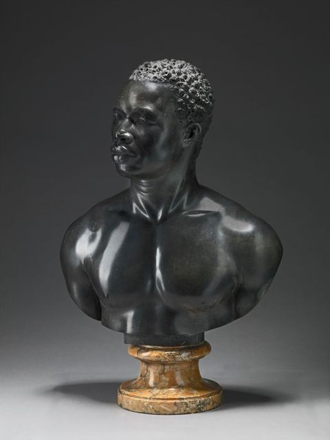 Sculpture. A bust includes the head, neck, and muscular chest and shoulders of a Black man. He has short, curly hair, noticeable eyebrow ridges, a wide nose, and his full lips are closed. Tendons stand out in in his neck, which turns halfway to look off to our left. Light gleams on the polished surfaces of the black limestome.