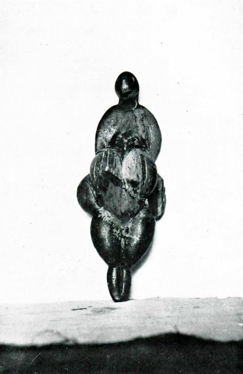 Sculpture. An angular carved statuette of a roughly female form. The form seems to have a small head over dramatically sloping shoulders. Heavy pendulous breasts hang over a round belly and tapering legs.