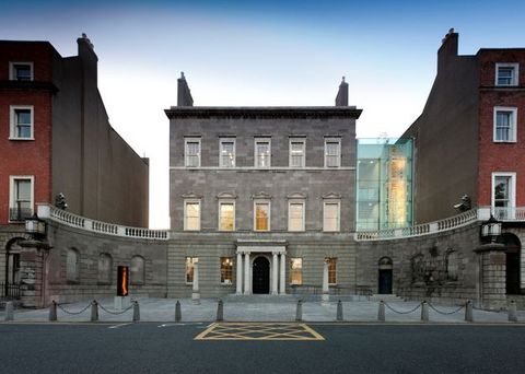 Architecture. A three-storey, square, grey stone building sits between two red brick buildings. Wings extend from the ground level curving towards us, like arms. A glass enclosure connects the grey building to the brick building on our right. On the other side, a gap, as if made by an alleyway, separates the grey structure from the other brick structure.