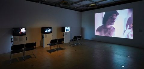 Installation. In a darkened room, a film is projected onto the wall to our right. Three television sets are on three stands along the wall to our left. Two chairs sit in front of each pedestal.