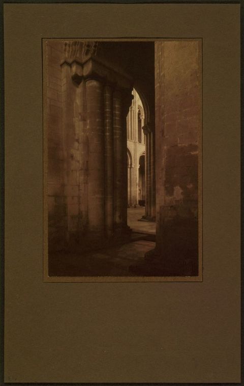 Sepia toned photograph. In tones of parchment and tawny brown, we look through a sliver of an opening into a light-filled nave of a cathedral. There are columns and rounded arches in both spaces.
