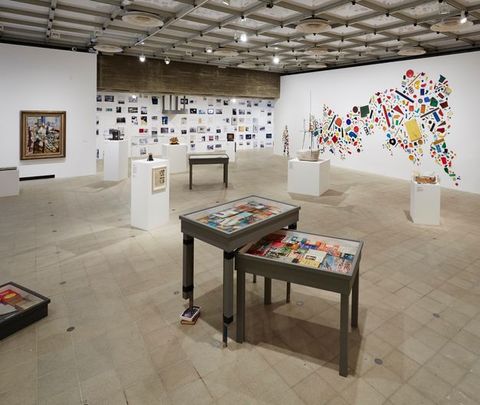 Installation. Tables or blocky podiums in a large, open space are freely placed so they do not form a pattern. Framed art, pictures, and possibly printed material are on the walls across from us. To our right, brightly coloured objects are affixed to a long wall to create the silhouette of a person and a large abstract form.