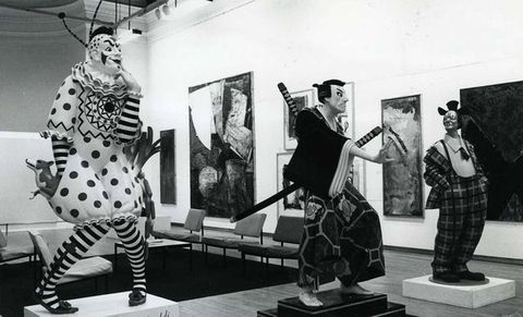 Installation photograph. This black and white photograph shows two life-sized clowns flanking a samurai. Each sculpture stands on a pedestal in a gallery hung with paintings or other media.