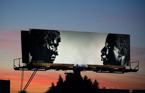 Photograph. A billboard is illuminated against a sunset sky of coral red, deep pink, and vivid blue. On the billboard are two black stone busts facing each other from either end of the long composition. To our left, Antico’s sculpture shows a person with full, curly hair, a straight nose, and defined cheekbones. His chin is slightly tucked in. To our right, the face of Harwood’s sculpture seems to look levelly back. It depicts a Black man with short, curly hair, a softly curved nose, and full lips which are closed. 