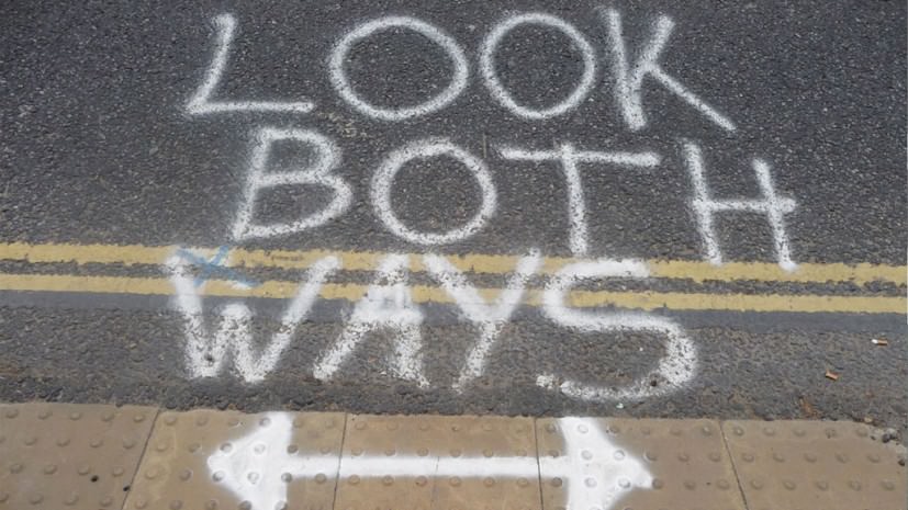 Graffiti art. A street is spray-painted in white with the words 'Look both ways' above a double-sided arrow. Double yellow lines cross the picture, and textured pavers indicate that this is where a sidewalk meets the street.