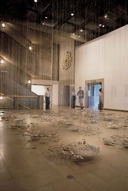 Installation photograph. Water appears to fall from a dark panelled ceiling to the floor, onto twenty-five works that seem to be made of natural materials and bits of glinting metal. Four people across the room look at the installation.