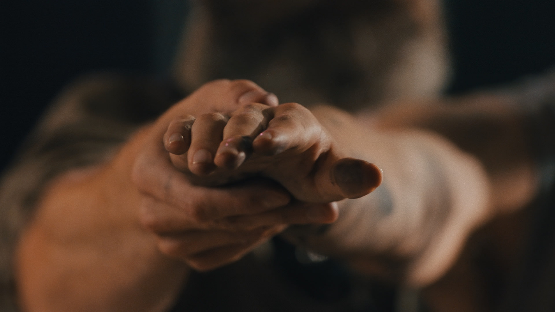 Video still. Sharp in focus, two pale-skinned hand come towards us. One hand is flat, palm down, and the other cups around the pinkie edge side. The arms extend away, out of focus, towards a dark background.