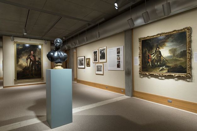 Installation photograph. Harwood's sculpture Bust of a Man, in polished black limestone, sits facing us on a tall pedestal. A partial wall beyond the bust is hung with a painting of a pale-skinned man wearing armour, attended by a boy with brown skin. A wall to our right is hung with a painting of a pale-skinned man wearing a black hat and green coat, with a rifle hung over his arm, attended by a boy or young man with brown skin in a bright red tunic and dark jacket. They are in a landscape with trees.