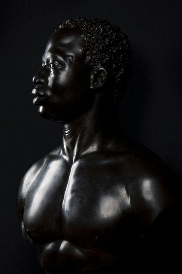 Photograph. Edges of a sculpture disappear against a deep black background. The sculpture faces our left in profile and nearly fills the picture.