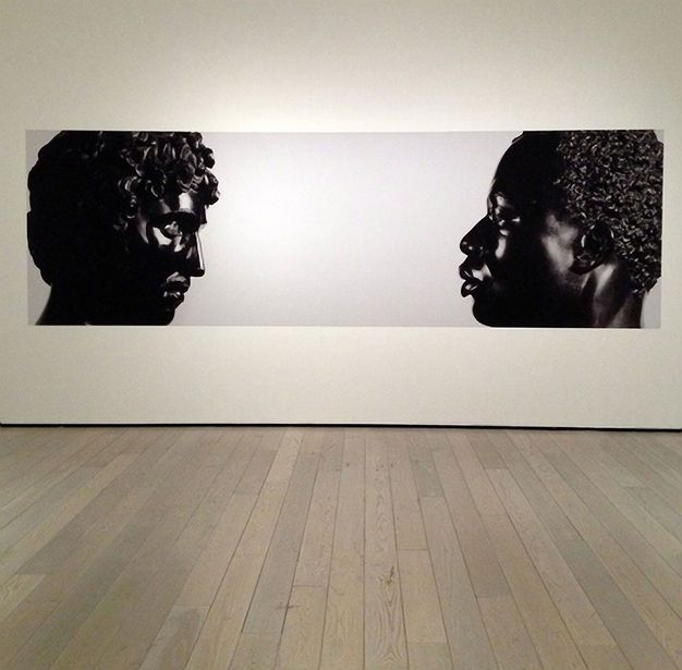 Photograph. From opposite ends of a long composition, two black stone busts face each other. To our left, Antico’s sculpture shows a person with full, curly hair, a straight nose, a moustache, and defined cheekbones. His chin is slightly tucked in. To our right, the face of Harwood’s sculpture seems to look levelly back. It depicts a Black man with short, curly hair, a softly curved nose, and full lips which are closed. Both are shown to the tops of their shoulders.