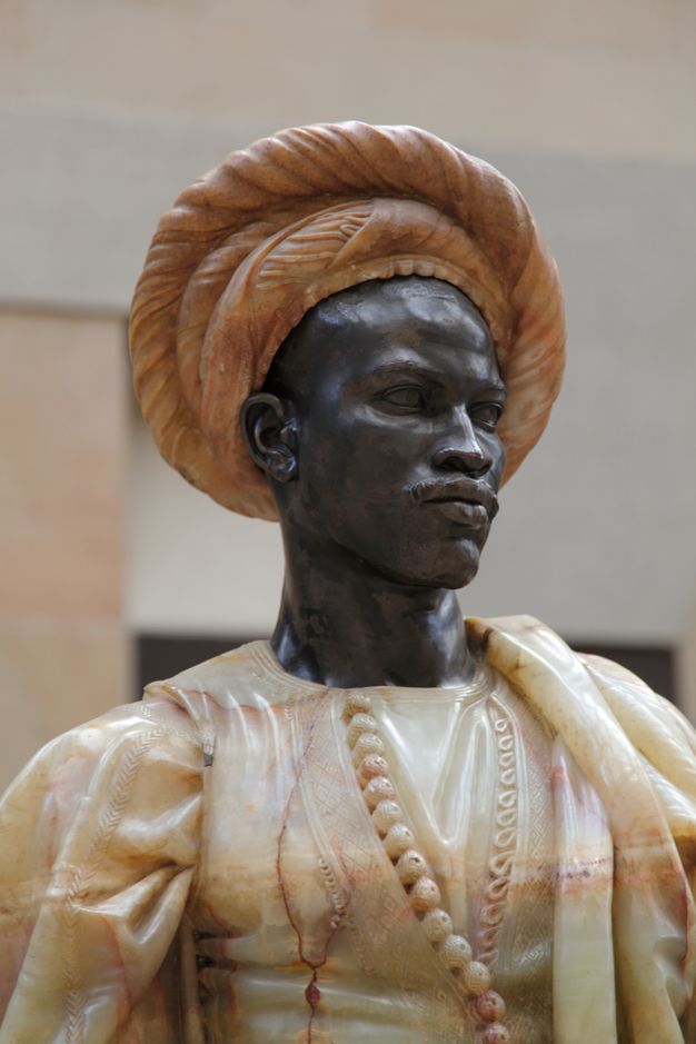 Sculpture. A bust includes the head, neck, and muscular chest and shoulders of a Black man. He has short, curly hair, noticeable eyebrow ridges, a wide nose, and his full lips are closed. Tendons stand out in in his neck, which turns halfway to look off to our left. Light gleams on the polished surfaces of the black limestome.