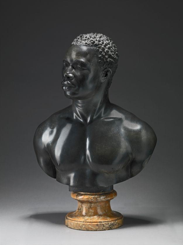 Sculpture. A bust includes the head, neck, and muscular chest and shoulders of a Black man. He has short, curly hair, noticeable eyebrow ridges, a wide nose, and his full lips are closed. Tendons stand out in in his neck, which turns halfway to look off to our left. Light gleams on the polished surfaces of the black limestome.