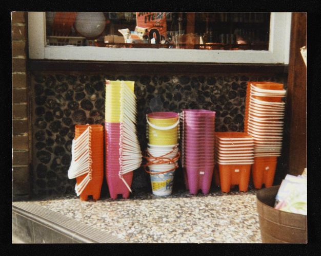 Colour photograph. Buckets and moulds for making sandcastles are stacked in six columns. Most of the moulds and buckets are orange, pink, or yellow, all with white handles. Some of the buckets are printed with what could be a landscape scene, and those have orange handles. They seem to be collected on a countertop.