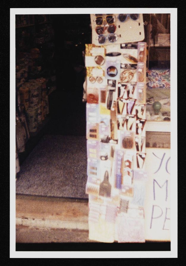 Colour photograph. A blurry view of a display showing sunglasses, hair clips, and combs. The edge of a handwritten sign with purple letters is to our right. A shadowy shop opens beyond the display, to our left.