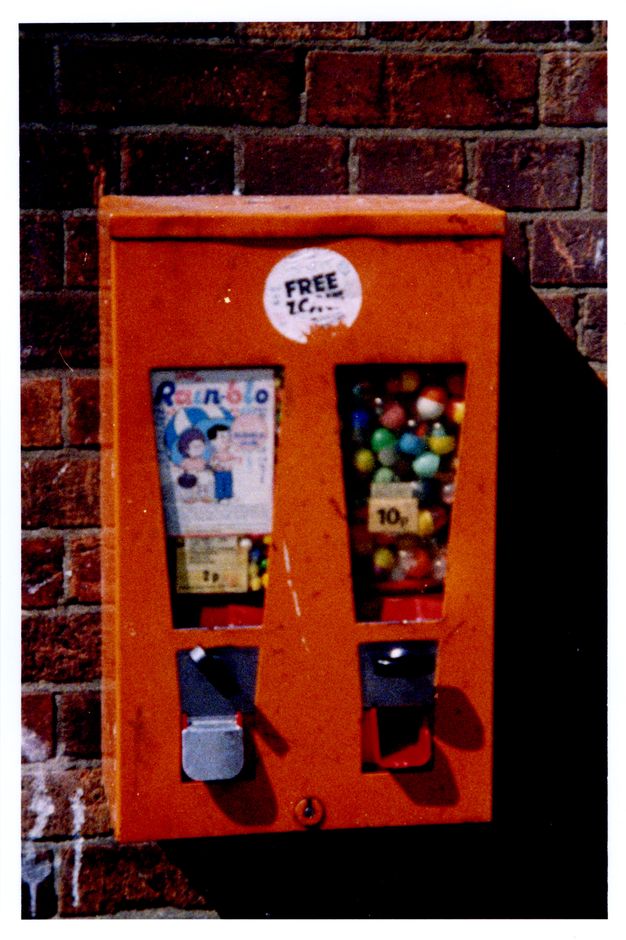 Colour photograph. Out of focus, a traffic-cone orange, double candy dispenser hangs on a brick wall. The candy to our left has a card reading 'Rain-blo' over two children standing under a striped umbrella. The dispenser to the right could sell bouncy balls or candy like jawbreakers. That window is labelled '10p'. A partially removed sticker at the top of the dispenser reads 'Free' over a word that has been torn away.