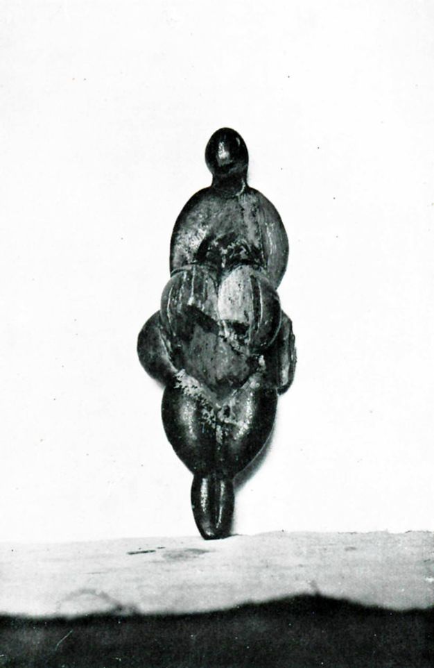 Sculpture. An angular carved statuette of a roughly female form. The form seems to have a small head over dramatically sloping shoulders. Heavy pendulous breasts hang over a round belly and tapering legs.
