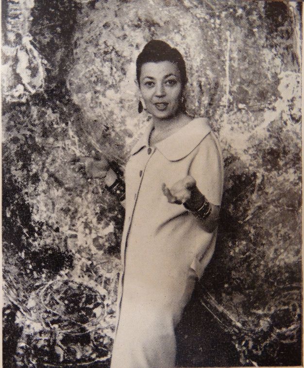 Black and white photograph. A woman, the artist Magda Cordell, stands in front of her painting Figure (Woman). The painting fills the background. Magda’s long white dress or coat has a wide, rounded collar and buttons down the front. She holds both hands up, and her lips are parted, as if about to speak or smile.