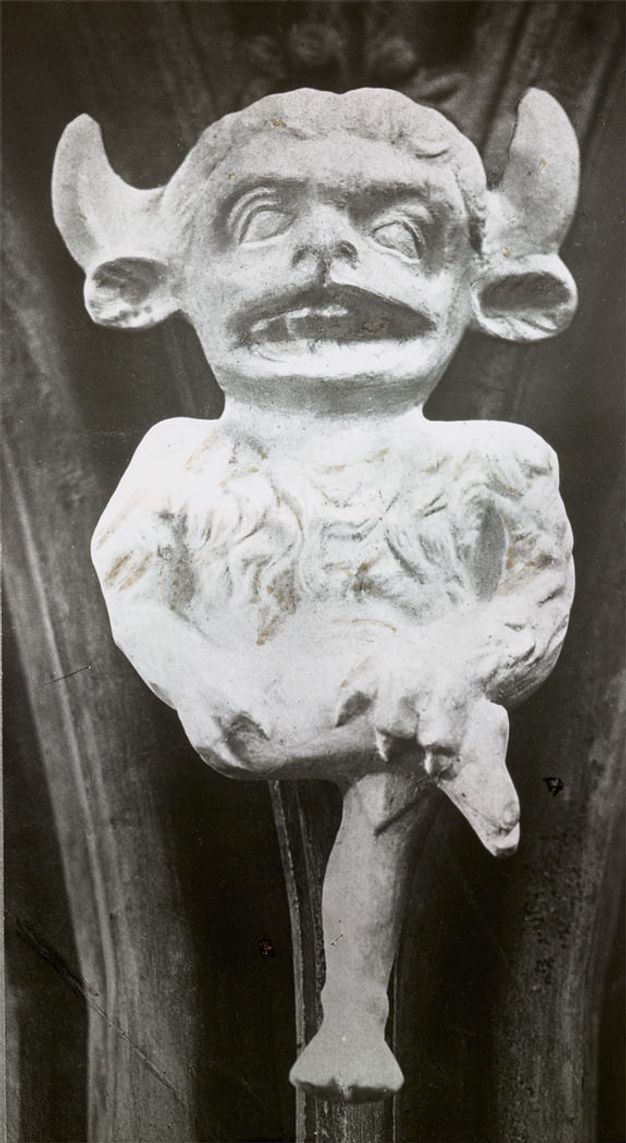 Black and white photograph. An imp has the legs of a person, a hairy torso and claws, perhaps of a lion. The head, which is larger in scale than the body, is a mix of animal and human features including lamb-like ears under horns. One leg is crossed over the other knee, and the mouth is open.
