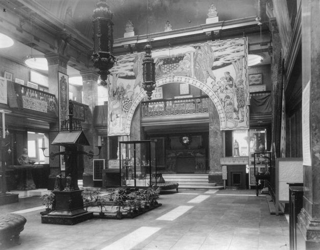 Black and white photograph. An open, hall-like space is hung with wall coverings, and furniture, plants, cases, and statuettes are arranged on the stone floor below.