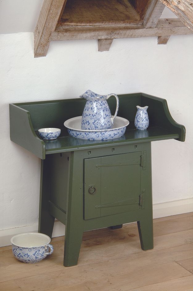 Furniture. This moss-green washstand has a cabinet base with four legs and a pull-ring door. The wide countertop has a high backsplash across the back and two sides. A pitcher in a basin sit in a hole in the counter next to a smaller bowl and pitcher. Those vessels, along with a chamber pot on the raw wood floor, are painted with a blue floral design.