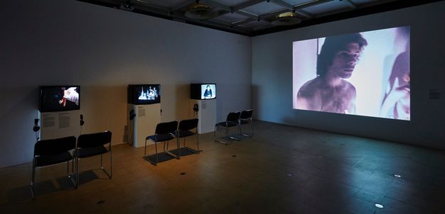 Installation. In a darkened room, a film is projected onto the wall to our right. Three television sets are on three stands along the wall to our left. Two chairs sit in front of each pedestal.