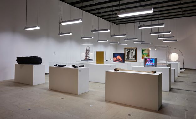 Installation. About a dozen long tables create rows across a long, white, nave-like room. Fluorescent lights hang down over the tables. Objects displayed on the tables include brooms, a pair of shoes, a television playing a video, and at least one photograph.