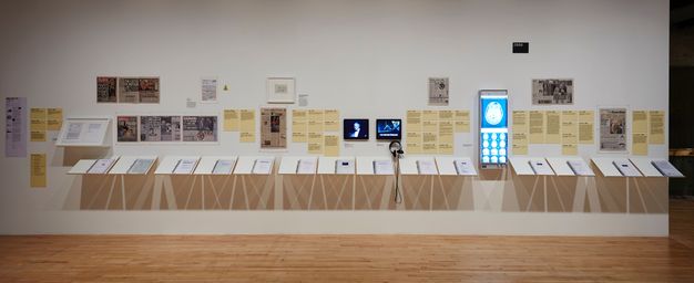Installation. Dozens of texts and pale yellow labels hang alongside clippings and other written material, two video screens, and a light box. Fourteen panels angle out from the wall below the texts.