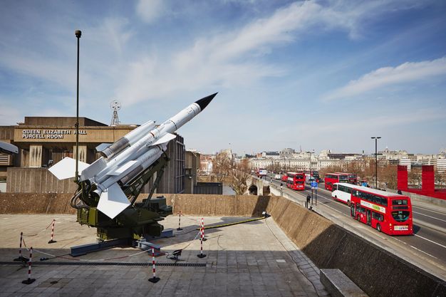 In front of a building identified on its front as 'Queen Elizabeth Hall Purcell Room', a missile launcher angles up at the sky. On a paved courtyard, the launcher is surrounded by red and white striped chains and stanchions. Several double-decker red buses drive on road running alongside the buildings to our right.
