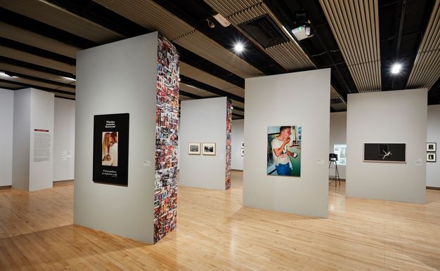 Installation. Four floating, block-like walls have one or two larger images on the side we can see. We can also see the sides of two blocks, though these surfaces angle sharply away from us. The latter are densely covered from floor to ceiling in smaller colour and black and white pictures.