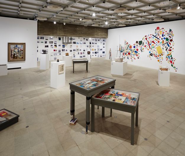 Installation. Tables or blocky podiums in a large, open space are freely placed so they do not form a pattern. Framed art, pictures, and possibly printed material are on the walls across from us. To our right, brightly coloured objects are affixed to a long wall to create the silhouette of a person and a large abstract form.