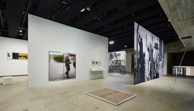 Installation. Shown on floating walls, photographs in colour and in black and white range from postcard to mural sized. The image we can see most clearly shows a soldier in camouflage holding a rifle. A board or upside-down frame lies on the floor in front of us.