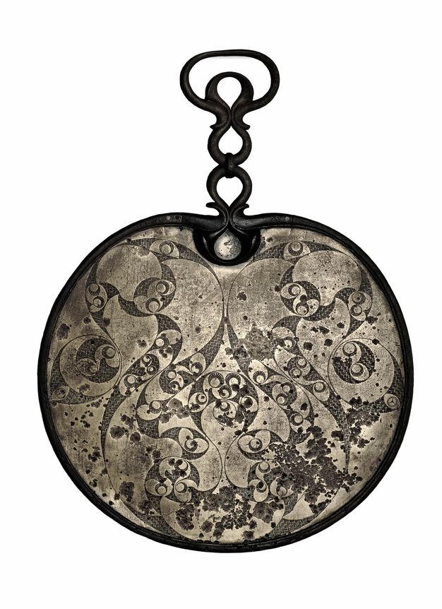 Mirror. The back of this bronze mirror is decorated with dark, swirling leaf-like and circular designs against a brighter background. A handle or hook at the top is made of three teardrop-shaped ornaments, the topmost set inside a larger oval.