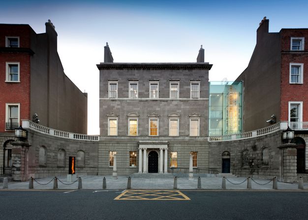 Architecture. A three-storey, square, grey stone building sits between two red brick buildings. Wings extend from the ground level curving towards us, like arms. A glass enclosure connects the grey building to the brick building on our right. On the other side, a gap, as if made by an alleyway, separates the grey structure from the other brick structure.
