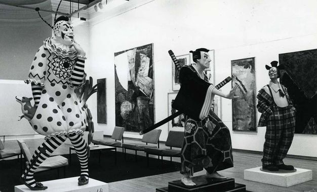 Installation photograph. This black and white photograph shows two life-sized clowns flanking a samurai. Each sculpture stands on a pedestal in a gallery hung with paintings or other media.