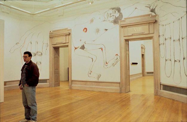 Installation photograph. An oversized line drawing of a nude man appears to have been created directly on the wall between two doors surrounded by tan-coloured moulding. He takes up the whole wall, his body, arms, and hands filling the space between and to either side of the doors. Touches of red scattered around his body suggest sores.