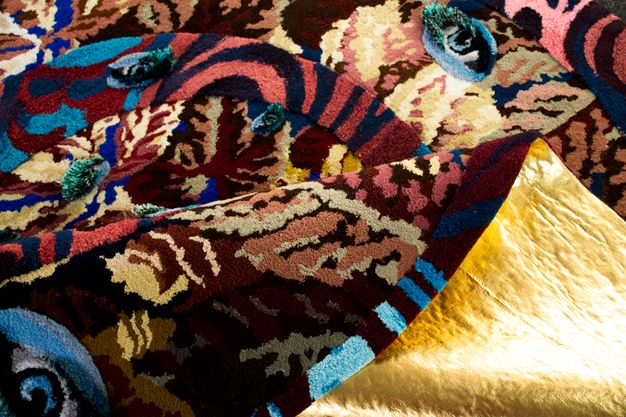 Tapestry. Patterns in maroon, dusky pink, white, navy, sky blue, sage green, and pale yellow intermix in this close view of low-pile rug. Several eyes of different sizes are woven among the patterns. The shimmering gold back of the rug is visible where it flips over near the bottom right corner.