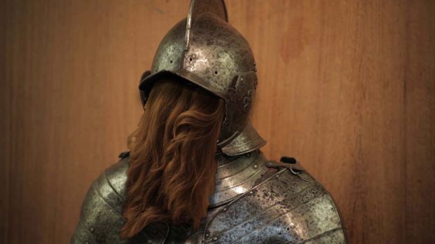 Video still. A suit of armour is shown from the shoulders up. Chestnut-brown, wavy hair pours from the opening where the face should be. The surface of the armour is darkened in areas, perhaps pitted.
