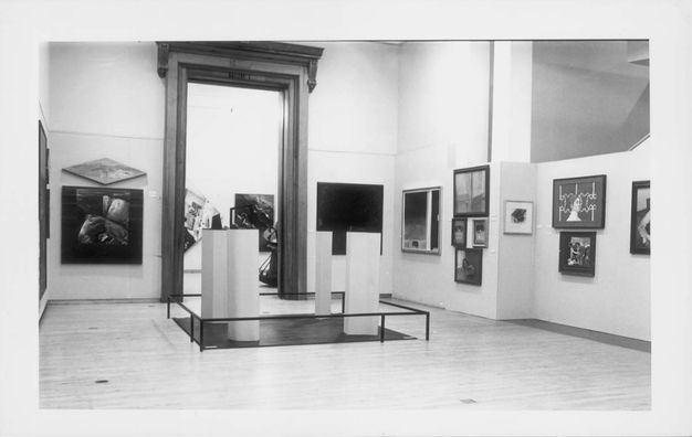 Installation photograph. Rectangular, square, or diamond-shaped works of art hang on the walls of a gallery. Four pillars stand enclosed by a low railing near the centre of the room.