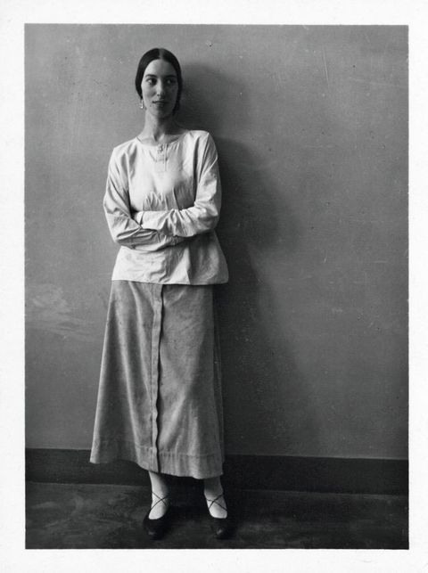 Black and white photograph. A woman stands against a wall with her arms folded across her body, looking off to our right. She has an oval face framed by black hair parted down the middle and pulled back. She wears a loose, long-sleeved shirt and a long skirt.