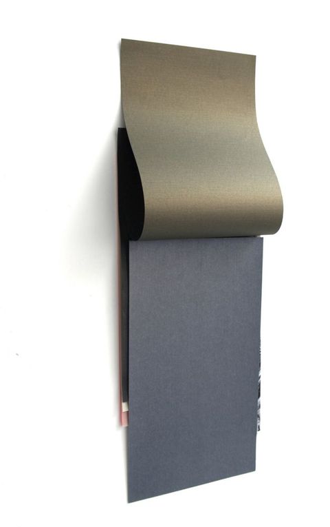 Installation photograph. A charcoal-grey piece of paper hangs on a white wall. A burnished gold-coloured sheet hangs from the wall above the grey, as if the sheets were glued together along the top like a notepad. Slivers of black, white, and pink paper peek out from under the grey sheet.