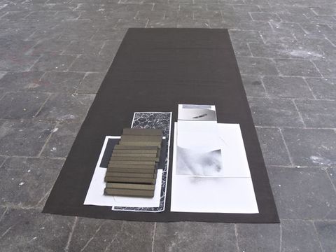 Installation photograph. Several works on paper are layered at one end of a long, rectangular black cloth laid on a grey stone floor. One sheet appears rather large, perhaps A2 size paper. Others are long strips or smaller pieces. Forms and patterns on the papers are in black, white, and shades of grey. Two golden brown pieces are folded like accordions.