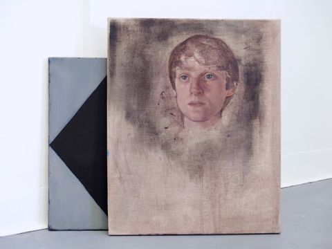 Installation photograph. Two works lean against the white wall of a gallery. The front painting is of a pale face gazing steadily off to our left with blue eyes. Straight brown hair falls in bangs and is cut close to the head. A dark grey area around the head is thinly applied, as if rubbed into the linen. The work tucked behind it is a design of grey and black triangles.
