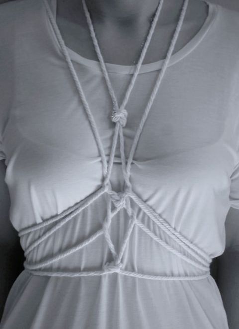Black and white photograph. A woman’s torso is tied with intricately knotted white rope over a white shirt. The contours of her bra are visible through the thin fabric.