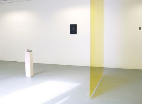 Installation photograph. A translucent yellow panel hovers over a grey floor, hanging perpendicularly to a white gallery wall. Two small works of art hang on the wide expanse of wall beyond the veil. A white, rectangular pedestal holds a few objects, though details are difficult to make out in this photograph.