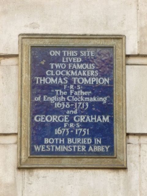 Plaque. A gold-framed sign on the outside of a building reads, “On this site lived two famous clockmakers Thomas Tompion F.R.S. ‘The Father of English Clockmaking’ 1638-1713 and George Graham F.R.S. 1673-1751 both buried in Westminster Abbey”.